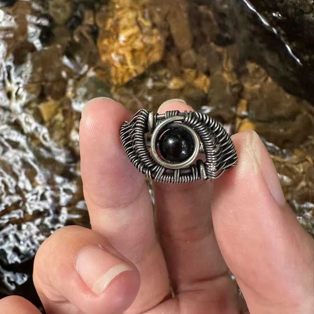The Eclipse natural black obsidian ring with hand-shaped brass wirework, held outdoors forscale