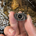 The Eclipse natural black obsidian ring with hand-shaped brass wirework, held outdoors forscale